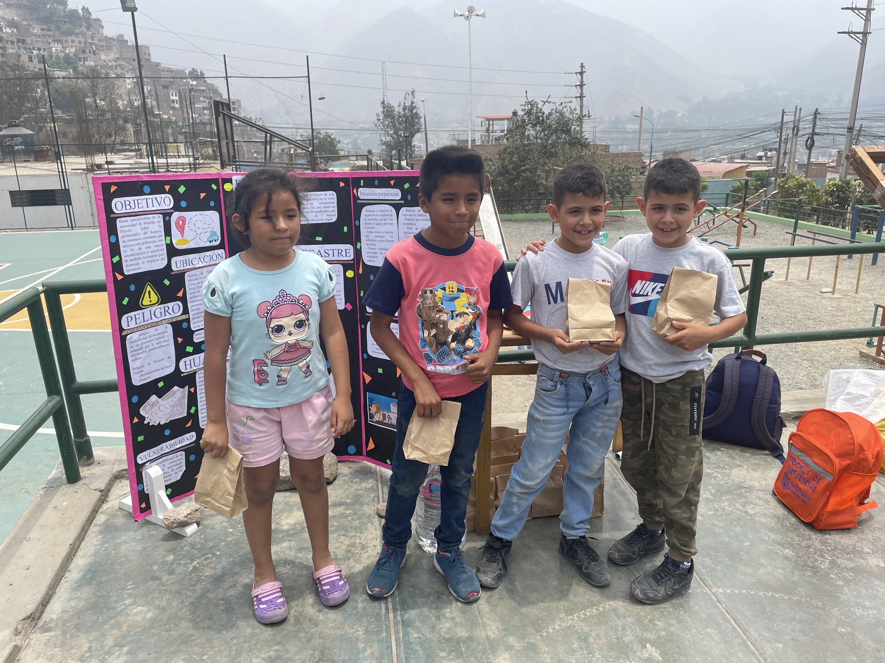 Gruppe von Kindern auf einem Basketballfeld mit Papierbeuteln in den H├Ąnden, mit Vorr├Ąten auf dem Boden und einer Tafel mit Text links, vor dem Hintergrund von Z├Ąunen, Masten, Stra├čenlaternen, B├Ąumen, Geb├Ąuden, H├Ąngen und Himmel.