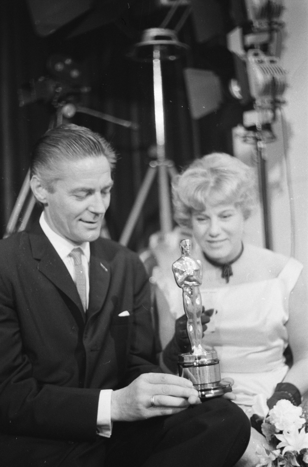 Ein Mann in einem schwarzen Blazer, einem weißen Hemd und einer Krawatte hölt eine Oscar-Statue, waehrend er neben einer Frau in Abendgarderobe sitzt, die einen Blumenstraußen hölt, mit weicher Beleuchtung im Hintergrund.