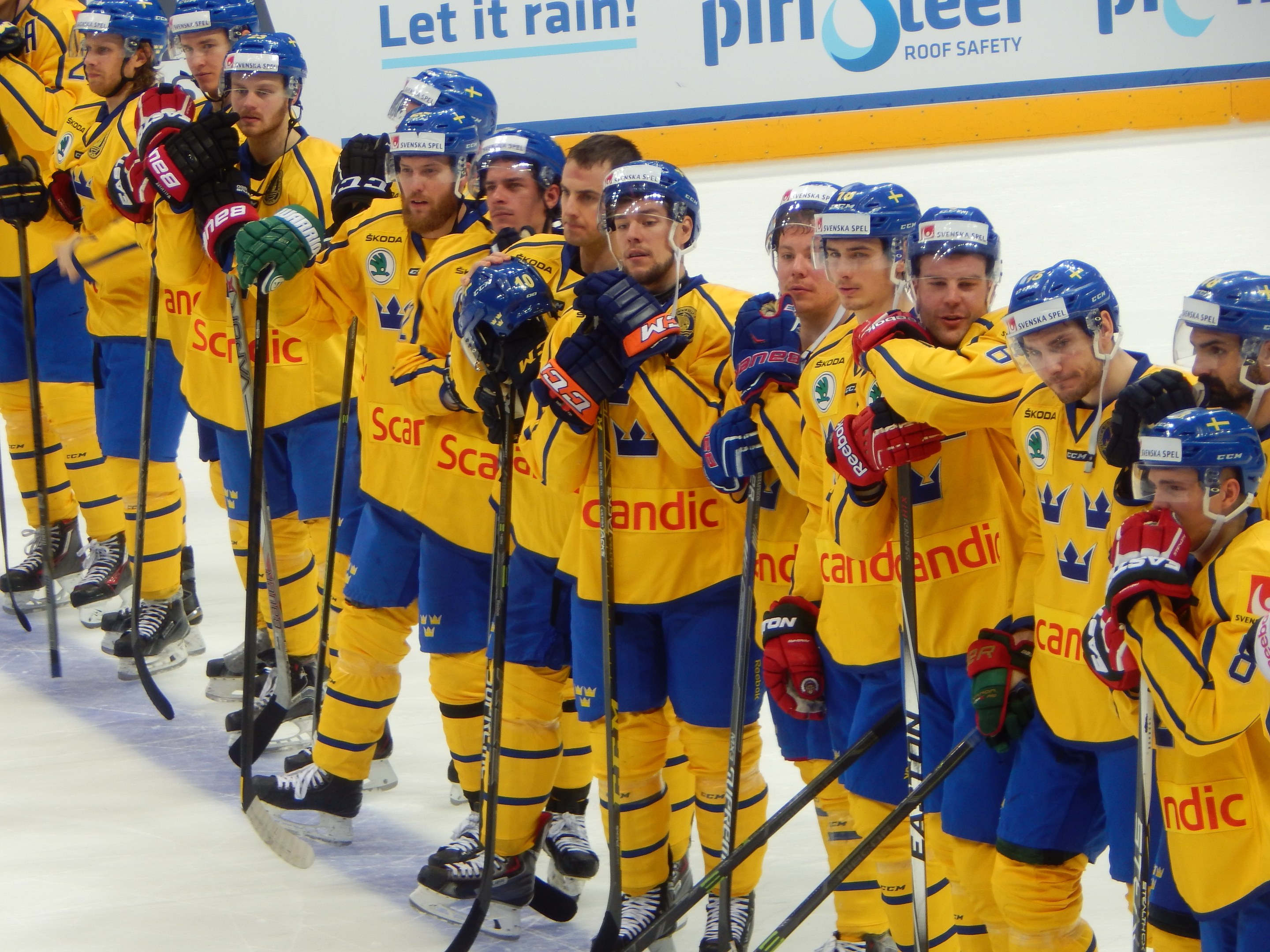 Eine Gruppe von Männern in gelb-blauen Eishockeyuniformen steht auf einem Eisstadion und hält Hockeystöcke, mit einer Tafel im Hintergrund.