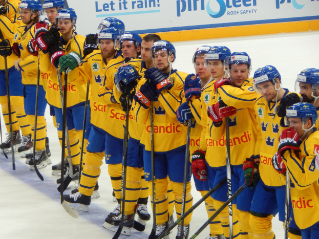 Eine Gruppe von Männern in gelb-blauen Eishockeyuniformen steht auf einem Eisstadion und hält Hockeystöcke, mit einer Tafel im Hintergrund.