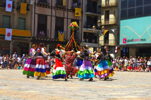 Eine Gruppe von Frauen in bunten Kleidern, die auf einer Straße tanzen, umgeben von einer sitzenden und stehenden Menge, mit Gebäuden, Fahnen, einem Laternenpfahl und einem Schild im Hintergrund.