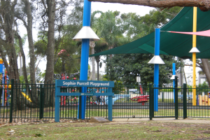 Ein Spielplatz mit grüner Überdachung, Metallzaun, Namensschild, Pfosten, Bäumen, spielenden Kindern, Gebäuden und einem bewölkten Himmel.