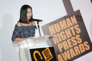 Eine Frau in einem schwarzen Kleid steht an einem Podium mit einem Mikrofon und hält eine Rede bei den Human Rights Press Awards, mit einem ernsten und konzentrierten Gesichtsausdruck.
