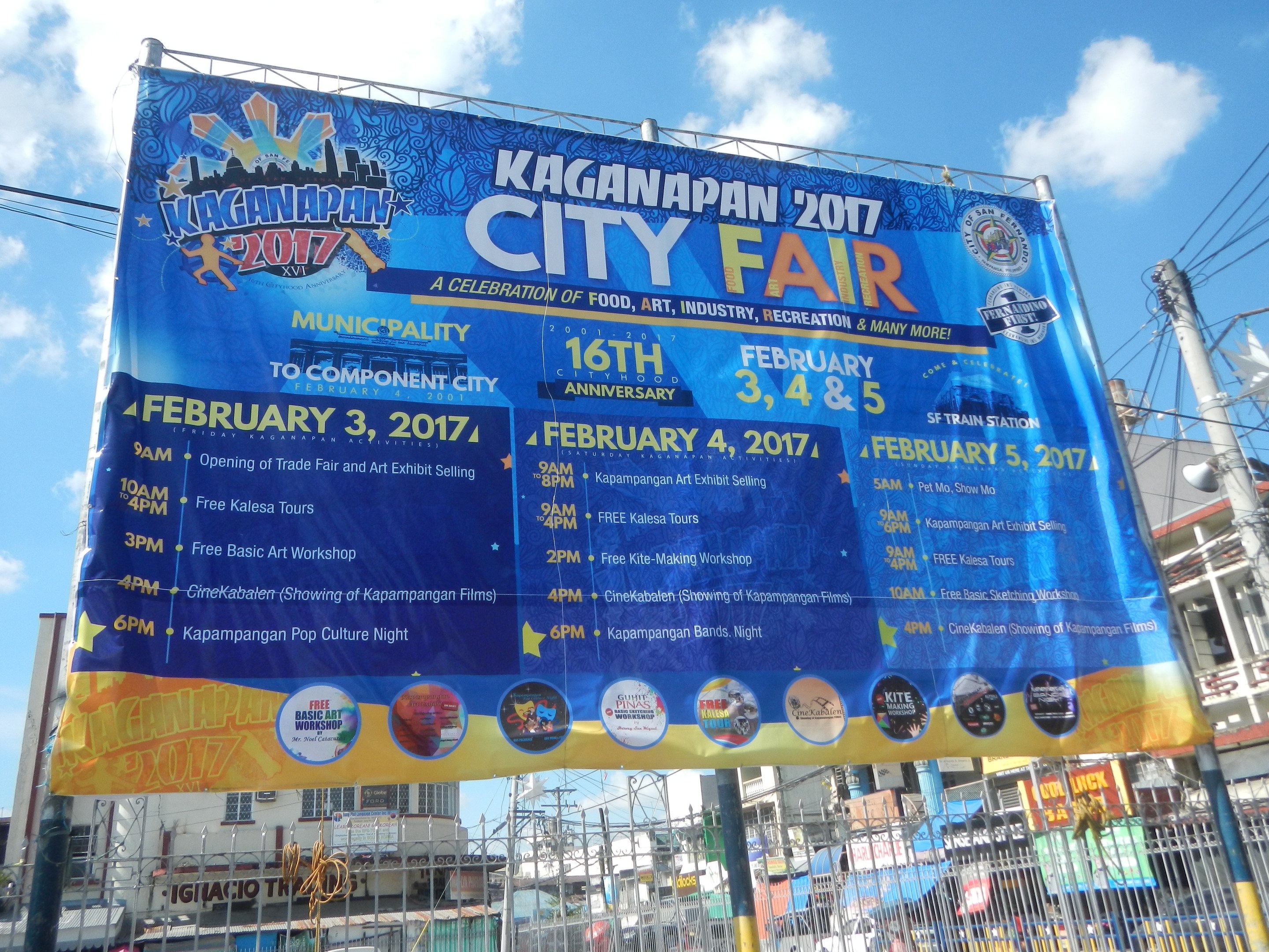Plakat mit der Aufschrift 'Kaganapan City Fair 2017' umgeben von städtischen Elementen unter einem klaren blauen Himmel.