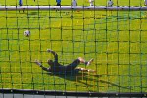 Fußballspieler hält vor dem Tor, Netz im Vordergrund und einige Personen im Hintergrund sowie mehrere Bälle auf dem Boden verstreut.