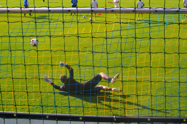 Fußballspieler hält vor dem Tor, Netz im Vordergrund und einige Personen im Hintergrund sowie mehrere Bälle auf dem Boden verstreut.