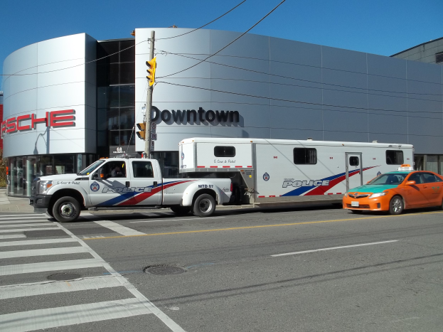Ein Polizeiwagen vor einem Gebäude mit Text geparkt, eine Ampel mit einem Mast und Drähten im Vordergrund und Bäume mit einem klaren blauen Himmel im Hintergrund.