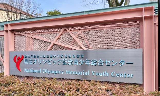 Außenansicht des Nationalen Olympischen Memorial Youth Center mit einem Namensschild, Pflanzen am Boden, Bäumen und Gebäuden im Hintergrund und einem klaren blauen Himmel.