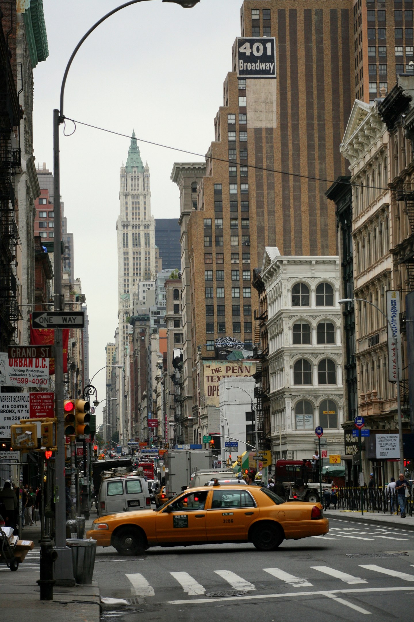 Gelbes Taxi fährt auf einer belebten Stadtstraße mit hohen Gebäuden auf beiden Seiten, Fußgängern auf dem Gehweg, Ampeln und Texttafeln, unter einem sichtbaren Himmel.