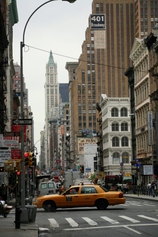 Gelbes Taxi fährt auf einer belebten Stadtstraße mit hohen Gebäuden auf beiden Seiten, Fußgängern auf dem Gehweg, Ampeln und Texttafeln, unter einem sichtbaren Himmel.