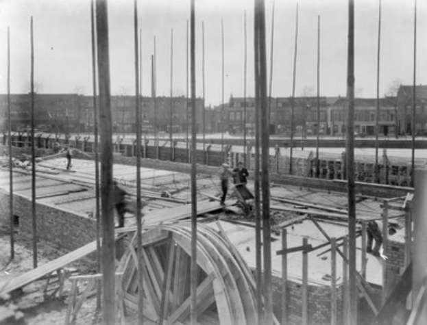 Schwarz-weiß-Foto einer Baustelle mit Arbeitern, hölzernen Pfählen und einem Rad im Vordergrund, Gebäuden, Bäumen und einem klaren Himmel im Hintergrund.