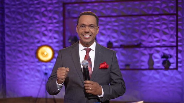 Stephen Colbert, Moderator von *The Daily Show with Trevor Noah*, steht auf einer Bühne und hält ein Mikrofon, trägt einen Anzug, Krawatte und Brille, mit einem Scheinwerfer und Ständer hinter ihm.
