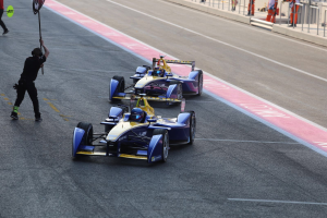 Zwei Formel-E-Autos auf einer Rennstrecke mit einem Fahne schwingenden Mann daneben, ein paar Leute links und Geländern mit mehr Leuten im Hintergrund.