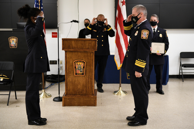 Gruppe uniformierter Personen in Masken vor einem Rednerpult mit Mikrofon, umgeben von Fahnen, Stühlen und Tischen, bei einer Vereidigung eines neuen Feuerwehrchefs, wobei eine Person ein Zertifikat hält.