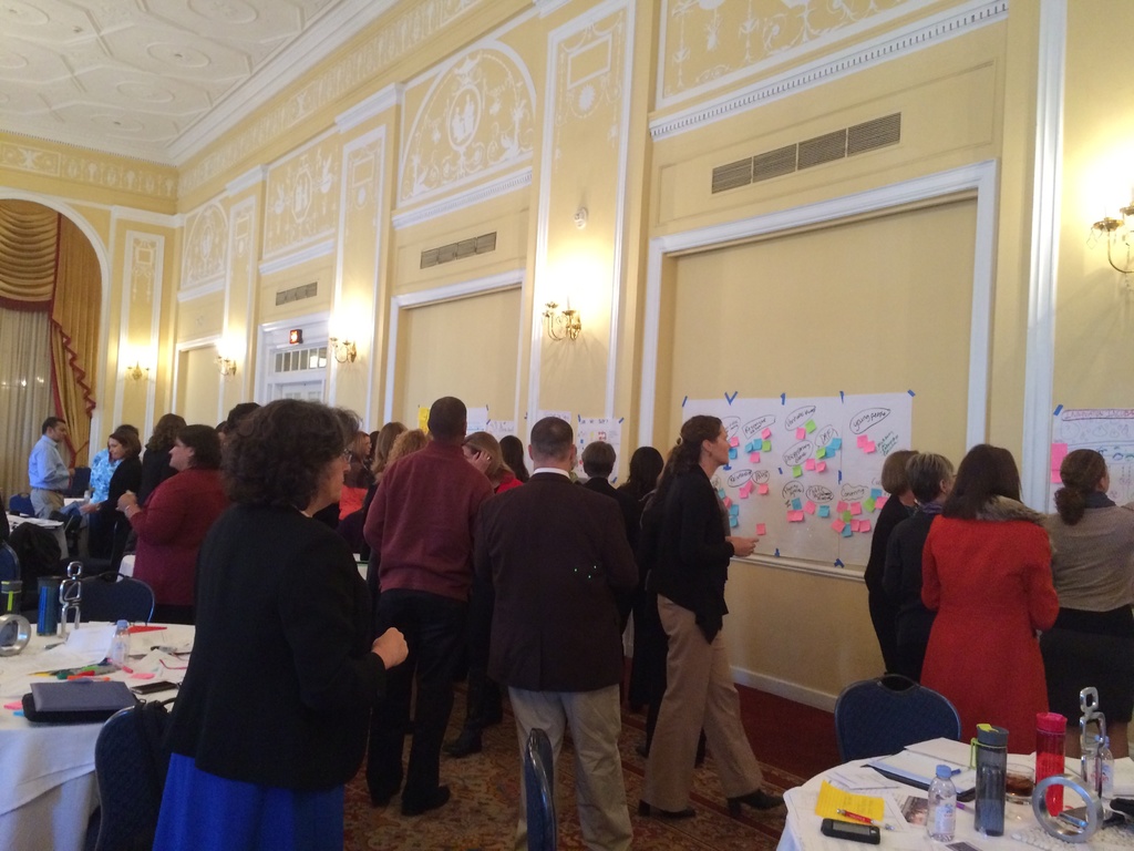 Eine Gruppe von Menschen untersucht Plakate und Diagramme, die an einer Wand in einem scheinbar Kunstmuseum angezeigt werden.