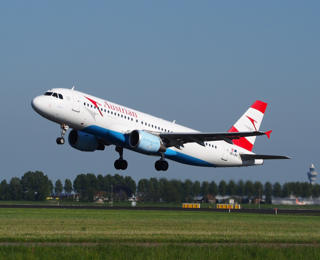Airbus A320-200 der Austrian Airlines startet vom Frankfurter Airport mit grünem Gras darunter, Bäumen und Gebäuden im Hintergrund und einem Turm auf der rechten Seite.