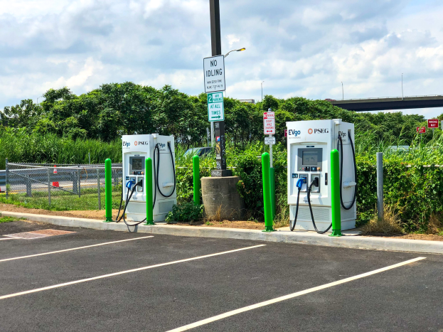 Elektrische Fahrzeug-Ladestation auf einem Parkplatz mit umgebenen Infrastruktur, Grünflächen und einer Brücke im Hintergrund bei einem bewölkten Himmel.