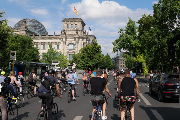 Eine Gruppe von Menschen, die auf Fahrrädern eine von Bäumen gesäumte Straße in Berlin, Deutschland, entlangfährt, mit Gebäuden und einer Bushaltestelle im Hintergrund, unter einem bewölkten Himmel und einer Flagge auf einem der Gebäude.