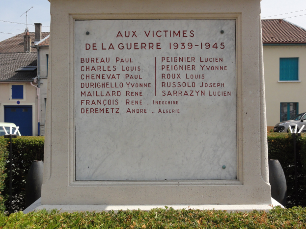 Ein steinernes Denkmal steht vor einem Gebäude mit einer Tafel, auf der "Aux Victimes de la Guerre 1939-1945" steht, umgeben von Grünfläche, parkenden Fahrzeugen, Versorgungsmasten und Wohnhäusern unter einem bewölkten Himmel.
