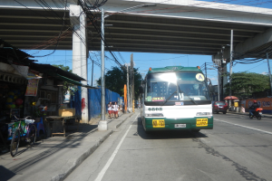 Ein grüner und weißer Bus fährt auf einer Straße neben einer Brücke, mit Strommasten und -leitungen entlang der Straße, Fußgängern auf dem Gehweg, parkenden Fahrrädern, Geschäften, Bäumen und einem klaren blauen Himmel im Hintergrund.