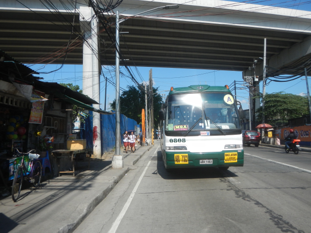Ein grüner und weißer Bus fährt auf einer Straße neben einer Brücke, mit Strommasten und -leitungen entlang der Straße, Fußgängern auf dem Gehweg, parkenden Fahrrädern, Geschäften, Bäumen und einem klaren blauen Himmel im Hintergrund.