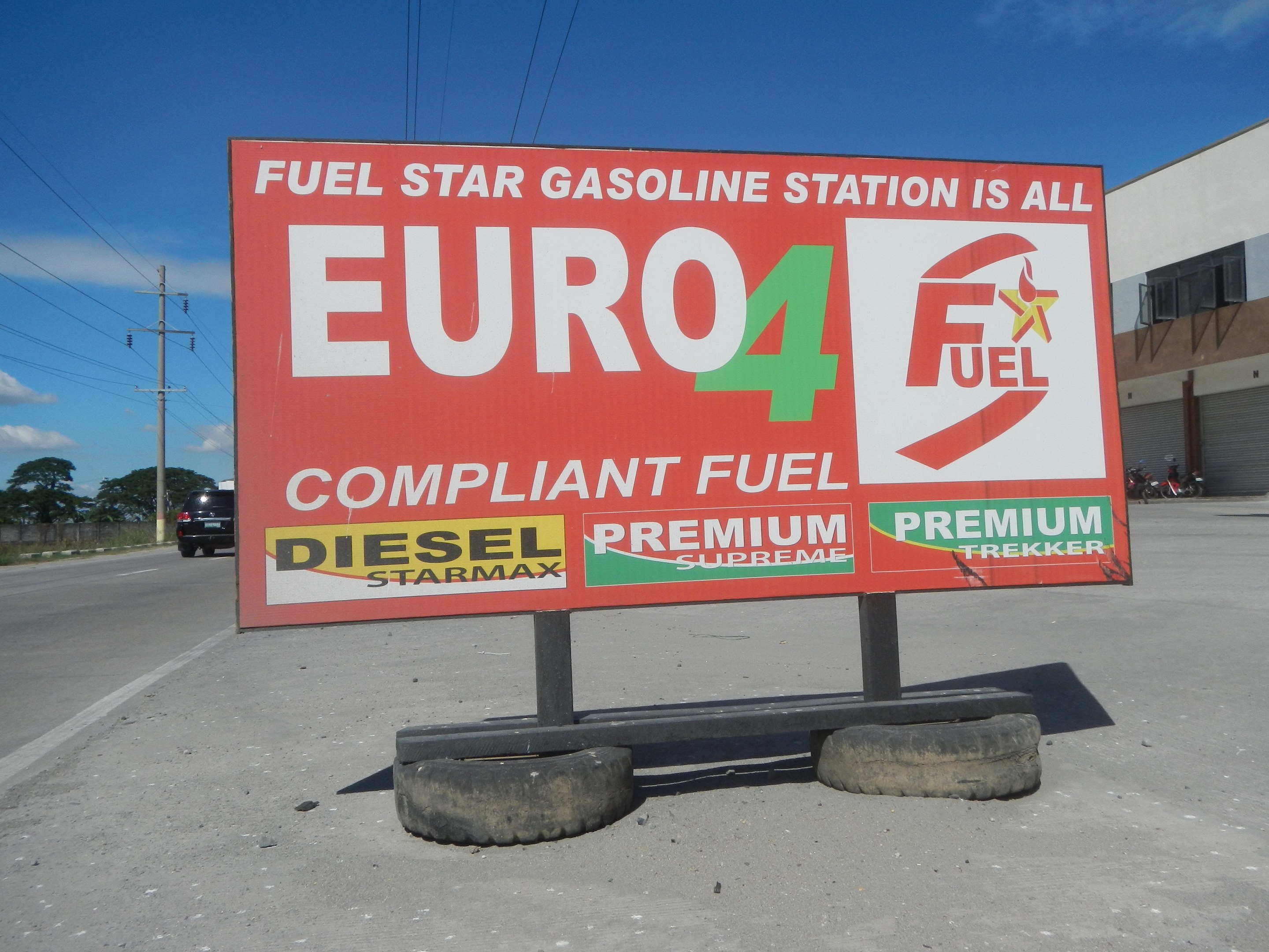 Eine Tankstelle mit einem "Euro4 Kraftstoff"-Schild im Vordergrund, Fahrzeuge auf der Straße, Strommasten mit Drähten, Bäume, ein Gebäude auf der rechten Seite und ein sichtbarer Himmel.