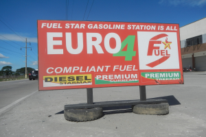 Eine Tankstelle mit einem "Euro4 Kraftstoff"-Schild im Vordergrund, Fahrzeuge auf der Straße, Strommasten mit Drähten, Bäume, ein Gebäude auf der rechten Seite und ein sichtbarer Himmel.