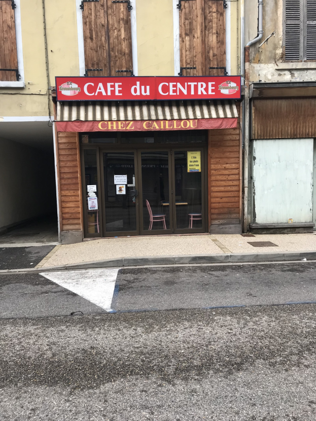 Ein zweistöckiges Gebäude mit einer 'Cafe du Centre'-Schrift auf der Seite, das Fenster, eine Tür und eine Straße davor mit einer kleinen Tafel in der Nähe zeigt.