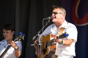 Ein Mann in einem weißen Kleid und mit Brille singt auf der Bühne in ein Mikrofon und spielt Gitarre, begleitet von einer Frau, die ebenfalls eine Gitarre spielt.