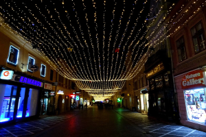 Eine nächtliche Stadtstraße mit Gebäuden auf beiden Seiten, die mit festlichen Weihnachtslichtern geschmückt sind, einige Menschen, die den Weg entlanggehen, und ein dunkler Hintergrund.