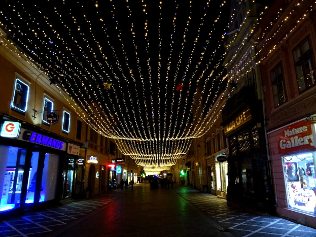 Eine nächtliche Stadtstraße mit Gebäuden auf beiden Seiten, die mit festlichen Weihnachtslichtern geschmückt sind, einige Menschen, die den Weg entlanggehen, und ein dunkler Hintergrund.