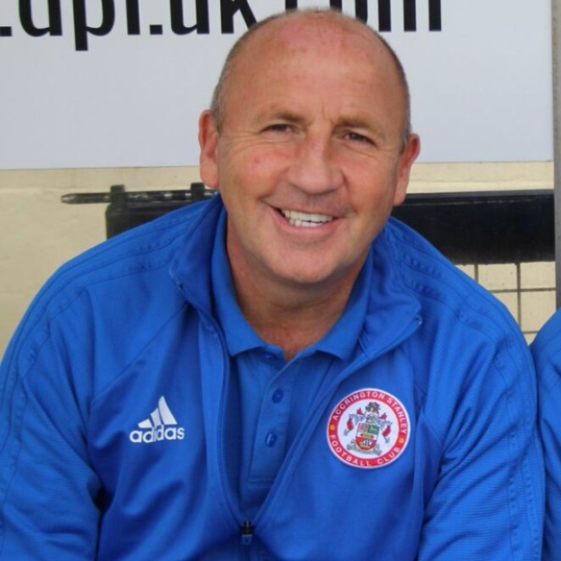Zwei strahlende Männer in einem Raum, einer in einem blauen Jackett, der wahrscheinlich als Trainer des Accrington Stanley Football Club identifiziert wurde, mit einer Tafel an der Wand dahinter.
