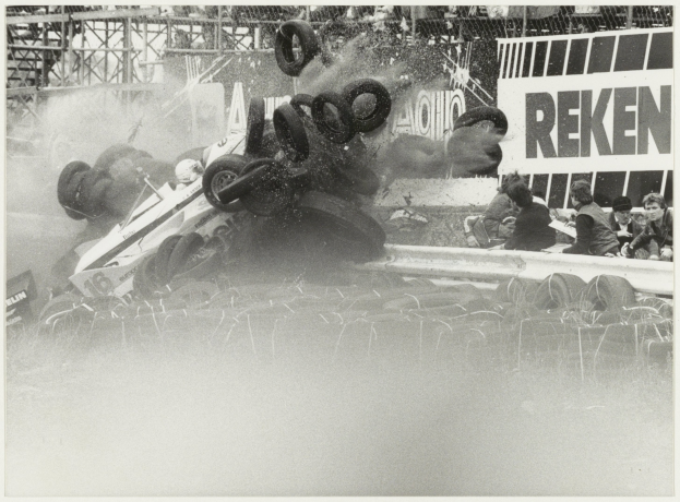 Schwarß-weißes Foto eines Rennwagens, der in einen Zaun gekracht ist und von Reifen umgeben ist, mit Menschen zu seiner Rechten und einer Tafel mit Text im Hintergrund.