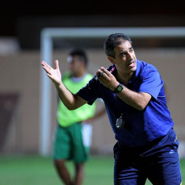Ein Fußballtrainer in einem blauen T-Shirt und Hose steht auf einem Feld und hält ein Mikrofon in der Hand, während er in Richtung der Spieler gestikuliert. Im Hintergrund ist ein Torpfosten und eine Person in einem grünen Kleid zu sehen.