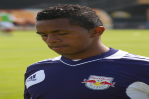 Ein junger Mann in einem blauen T-Shirt steht auf einem Fußballfeld, mit kurzem dunklen Haar und entschlossener Miene, unscharfer Hintergrund.