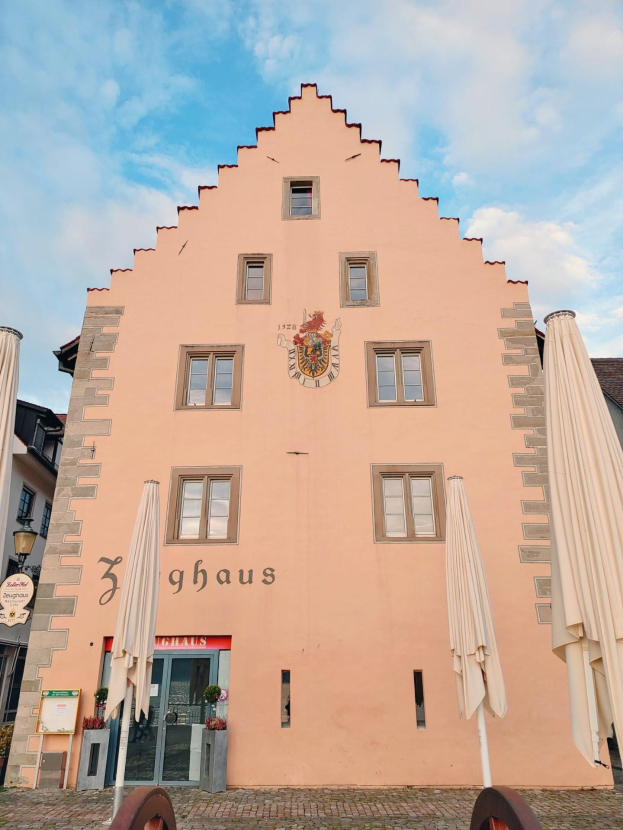 Ein pinkes Gebäude mit einem zentralen Uhrenturm, umgeben von Fenstern, Schirmen, einem Schild, einer Straßenlaterne, Topfpflanzen, unter einem bewölkten Himmel, repräsentiert München, Deutschland.