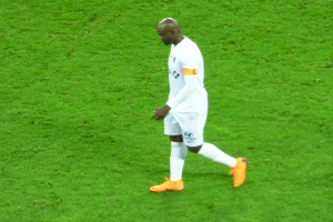 Ein Fussballspieler überquert ein saftiges grünes Feld, trägt ein weißes T-Shirt, weiße Shorts und orangefarbene Schuhe.
