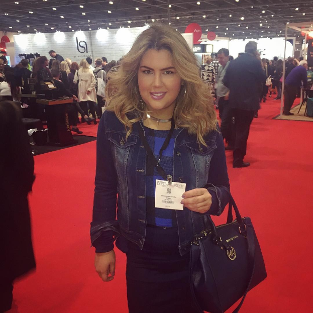 Eine Frau in Abendgarderobe steht auf einem roten Teppich bei einem Modeevent, lächelt und hält eine Handtasche, während Fotografen und Scheinwerfer im Hintergrund zu sehen sind.