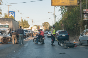Eine Gruppe von Menschen um ein verunglücktes Motorrad auf der Straße herumstehend mit mehreren Fahrzeugen, darunter ein Lastwagen, und einem Hintergrund aus Bäumen, Pfählen, Lampen und Schildern unter dem Himmel.
