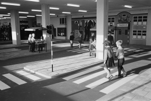 Ein Schwarz-Weiß-Foto von Kindern, die eine Straße an einer Ampel überqueren, mit Menschen, die stehen und sitzen, einer Ampel im Vordergrund und Säulen und Lampen im Hintergrund.